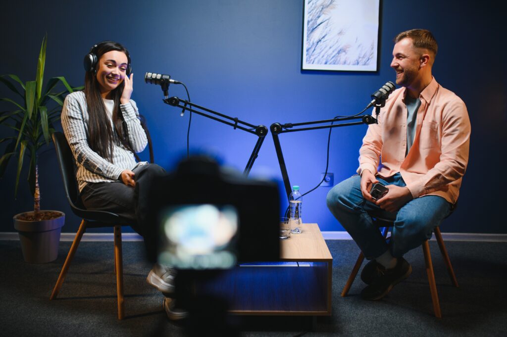 A woman and a man sit across from each other in a podcast studio, smiling and talking. Both have microphones on adjustable arms in front of them. The woman wears headphones and a striped shirt, while the man wears a light pink shirt and jeans. A camera is positioned in the foreground, recording the conversation.
